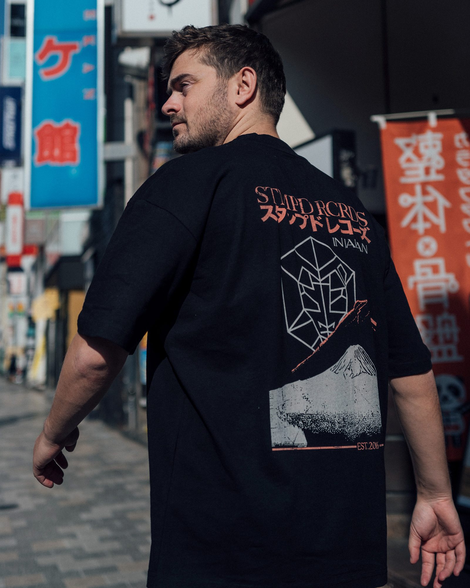 A person looking back over their shoulder in a shopping street with Japanese signs, in a black STMPD RCRDS T-shirt with the label's name in Latin and Japanese script, In Japan, its emblem and Mount Fuji printed on the back.