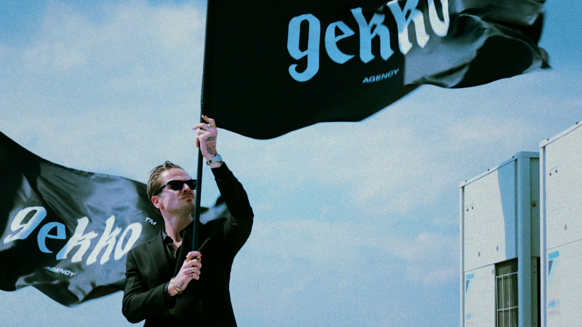 Still from the GEKKO Agency film: a person in sunglasses holding up a large black GEKKO Agency flag on a roof under a blue sky, a second flag beside them